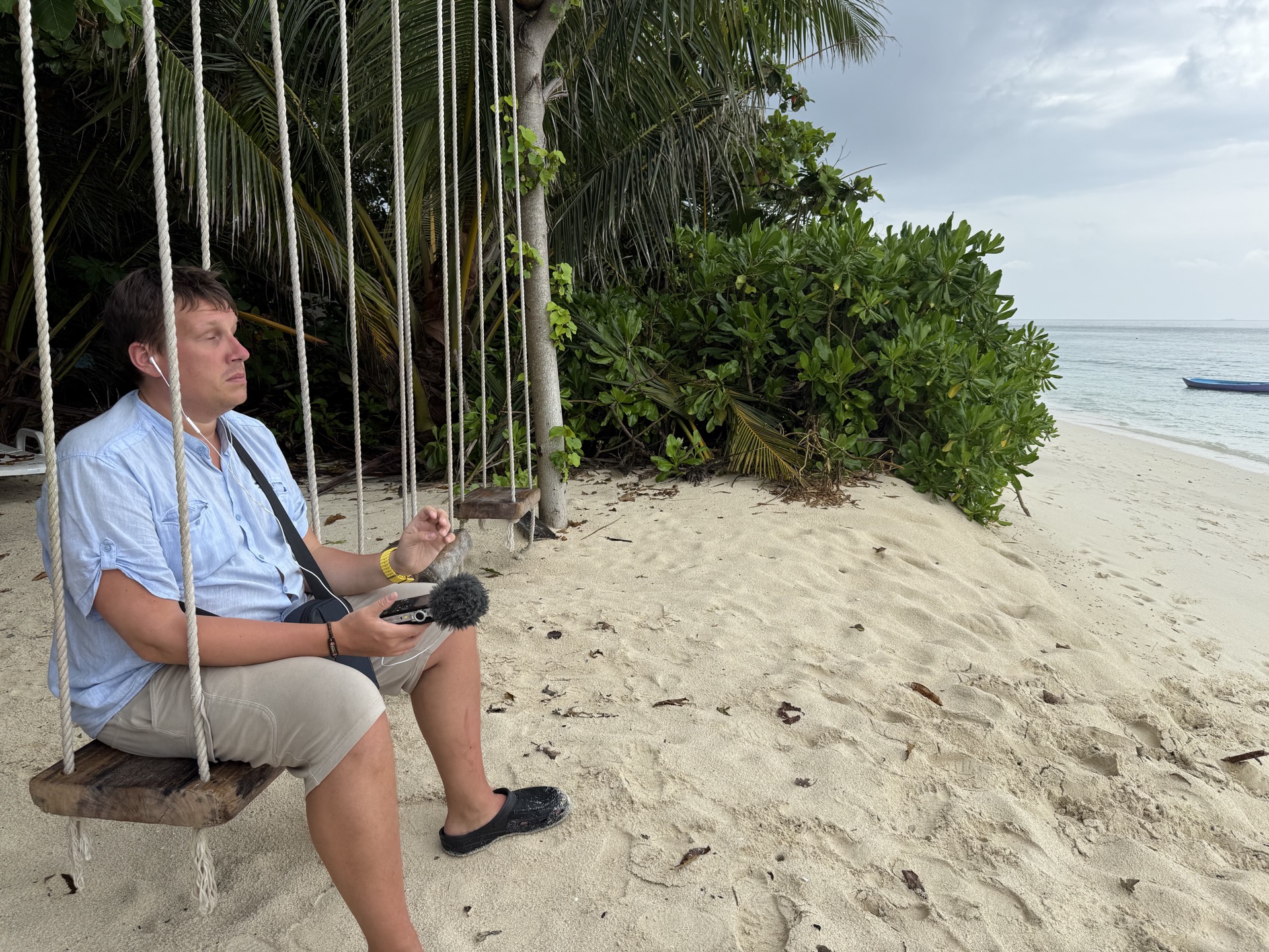 Jakob Rosin sitting on a rope swing on a white sand beach in the Maldives, holding a portable audio recorder with a furry windscreen microphone, earbuds in, gazing out at the calm ocean. Tropical vegetation and palm trees in the background.
